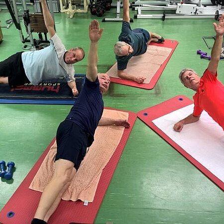 Vier Männer führen seitliche Plank-Übungen auf Yogamatten in einem Fitnessstudio aus, jeweils mit einem Arm nach oben.