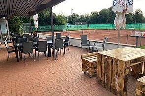 Terrasse eines Tennisclubs mit Holz- und Metallmöbeln, Blick auf rote Tennisplätze im Hintergrund.