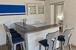 Moderne Küchenbar mit sechs blauen Hockern, über einem grauen Tresen, vor einer weißen Wand mit blauer Akzentlinie.