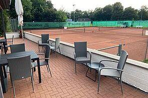 Terrasse mit Tisch und Stühlen, Blick auf einen Tennisplatz mit roten Sand und grünen Umrandungen.