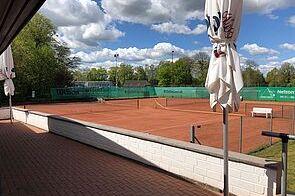 Tennisplätze mit rotem Sand und Sonnenschirmen unter wolkigem Himmel in einem grünen Umfeld.