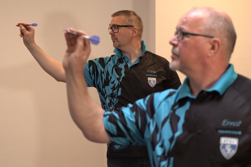 Zwei Männer in Dart-Shirts konzentrieren sich auf das Werfen von Darts in einem hellen Raum.