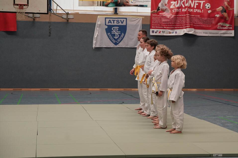 Gruppe von Judokas in weißen Anzügen mit gelben Gurten steht auf einer Matte in der Sporthalle, bereit für das Training.
