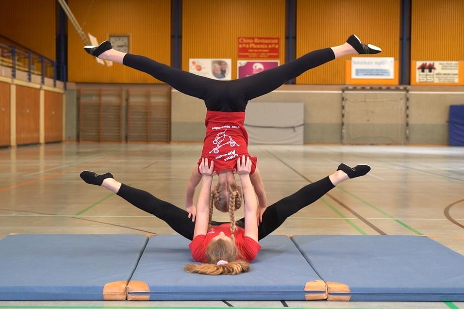 Zwei Akrobatinnen in roten T-Shirts führen eine Balanceübung auf einer Gymnastikmatte in einer Sporthalle aus.