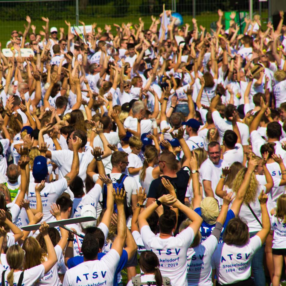 Große Menschenmenge mit weißen T-Shirts, die die Hände in die Luft heben, auf einem Freiluftgelände.