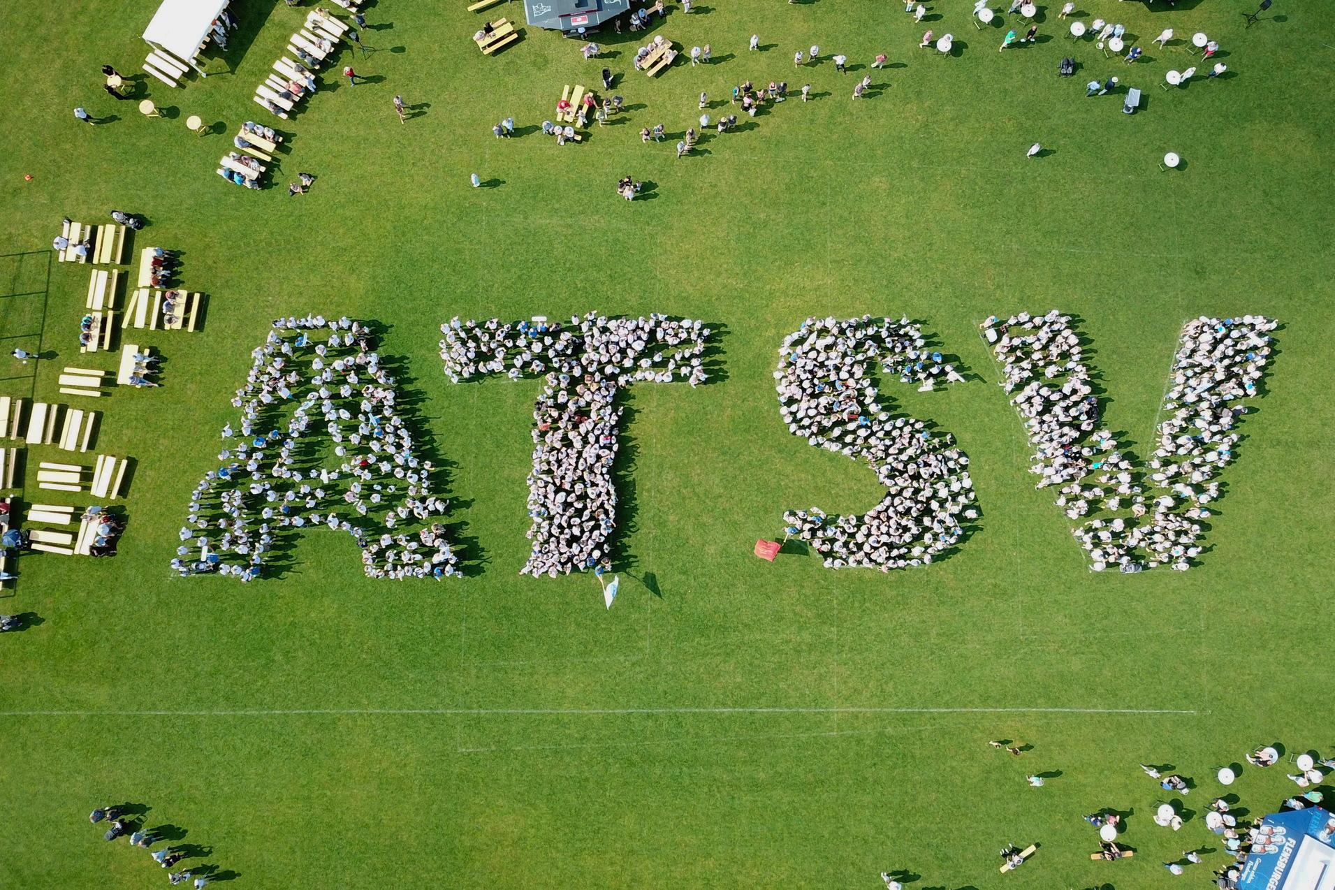 Vogelperspektive auf Menschen, die das Wort "ATSV" auf einer Rasenfläche bilden, umgeben von Tischen und Stühlen.