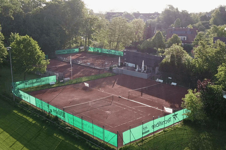 Luftaufnahme eines Tennisplatzes mit roter Erde, umgeben von Bäumen und einem Gebäude im Hintergrund.