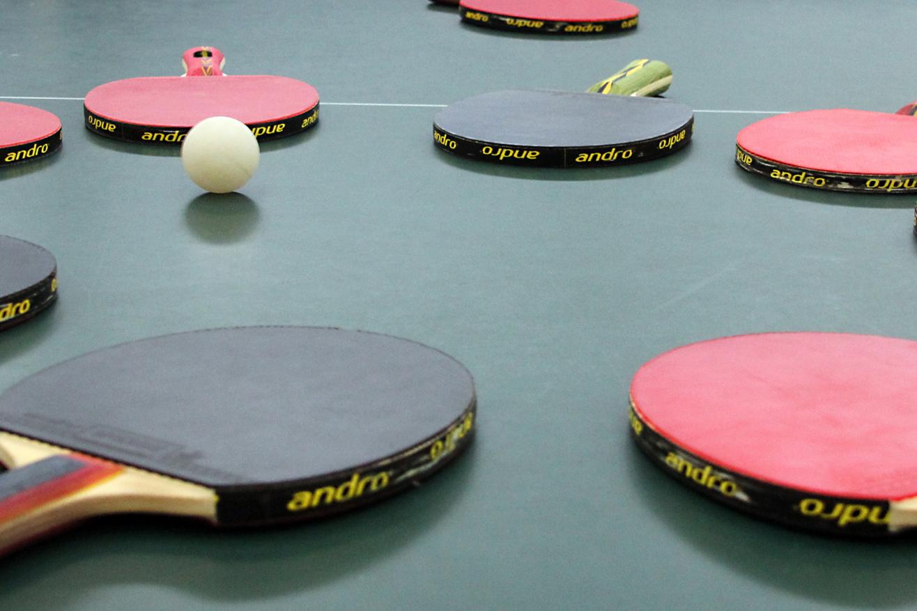 Mehrere Tischtennisschläger in verschiedenen Farben und ein Tischtennisball liegen auf einer grünen Tischtennisplatte.